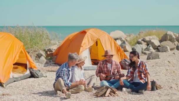 Tiro de cuerpo completo: dos mejores amigos parejas se sienta en la playa cerca de tiendas de campaña y divertirse juntos hablando, riendo y jugando ukelele — Vídeo de stock