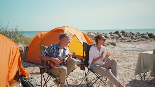 Hombre guapo camisa a cuadros está sentado en la silla del campamento con ukelele y canta canción divertida para su hermosa novia — Vídeo de stock