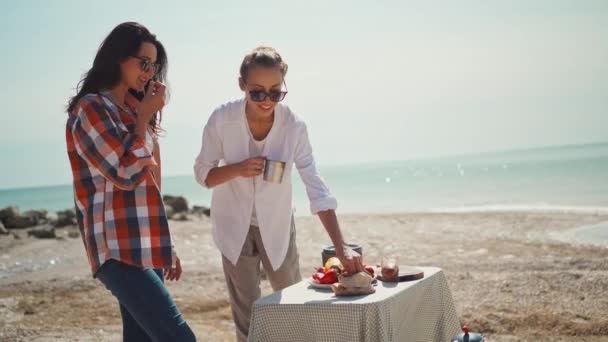 Δύο φίλες στέκονται στο τραπέζι της κατασκήνωσης με φαγητό για πικνίκ στην παραλία της θάλασσας, μιλώντας και τρώγοντας — Αρχείο Βίντεο