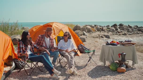 Młody dorosły mężczyzna w luźnym ubraniu przynosi cztery butelki piwa swoim przyjaciołom w obozowych krzesłach na plaży kemping z namiotami. — Wideo stockowe