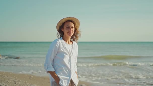 Atraktivní usmívající se žena cestovatel v bílé košili a slaměný klobouk chůze podél mořského pobřeží — Stock video