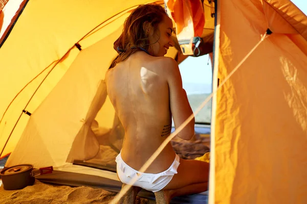 Solbrun jente med naken rygg som sitter i oransje telt under campingferien – stockfoto
