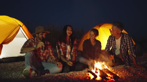 Grupo de amigos que se relajan por la noche en la playa acampando en tiendas de campaña, sentados en la hoguera con cerveza, hablando y disfrutando del calor del fuego — Vídeos de Stock