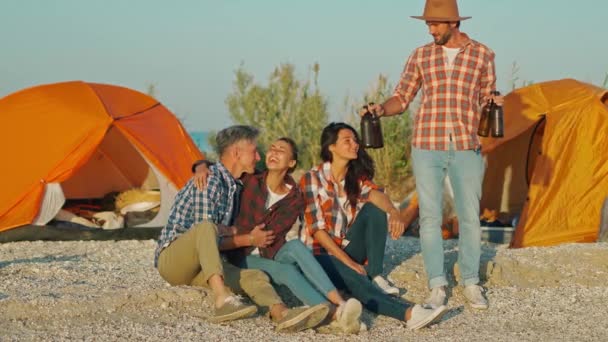 Guapo barbudo hombre adulto en ropa casual trae botellas de cerveza a amigos sentados en la playa de camping — Vídeos de Stock