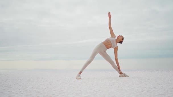 Parallaxe Shooting junge schöne fitte Frau steht in virabhadrasana yogische Haltung auf trockenen Salzsee Küste am bewölkten Morgen. — Stockvideo