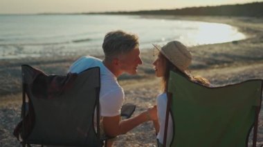 Arka manzara mutlu gezginler. Kamp sandalyelerinde oturan erkek ve kadın kumsalda güzel bir gün batımını öpüyor ve hayranlıkla seyrediyorlar.
