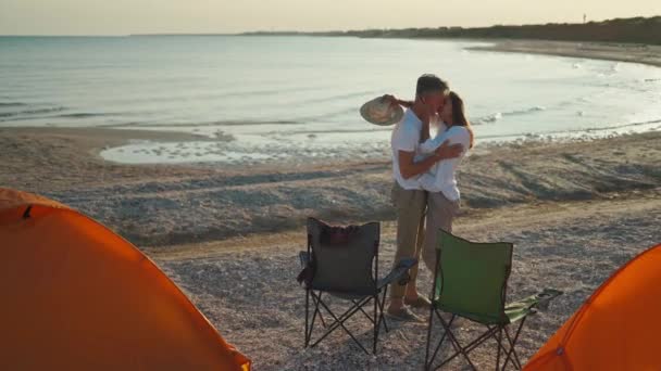 Felices viajeros enamorados hombre y mujer admiran hermosa puesta de sol en la playa. turistas junto a tiendas de campaña y sillas de campamento, abrazos, kissung, admirar el mar puesta de sol — Vídeo de stock