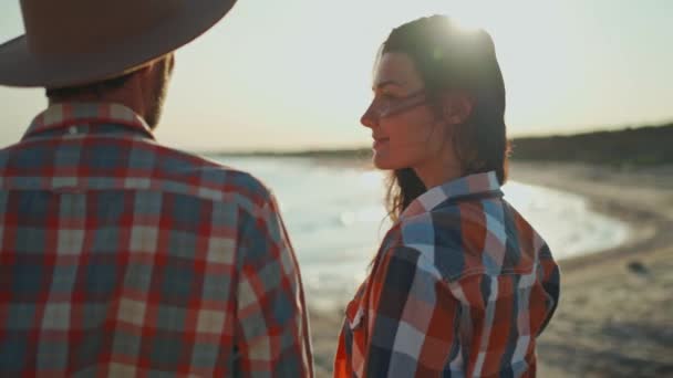 Ein Mann mit Hut und eine schöne asiatische Frau küssen sich am Strand bei Sonnenuntergang. schöne Paar Reisende in karierten Hemden bewundern schönen Sonnenuntergang am Meer. — Stockvideo