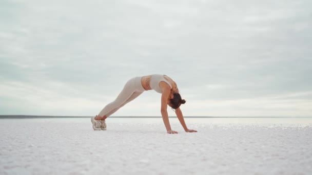 Самка практикует йогу на открытом воздухе, делая позы из последовательности йоги Sun Salutation на сухом берегу озера — стоковое видео