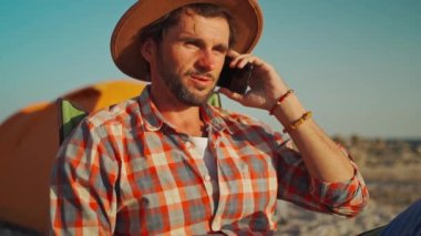 Şapkalı kaygısız iş adamı deniz kenarındaki çadırın yanında dinleniyor ve İş Arkadaşlarına Akıllı Telefon Çağrısı