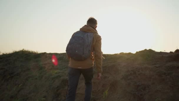 Camera follows man hiker with backpack climbs on steep hill at lake coast and looking sunrise at deserted sea beach — Stock Video