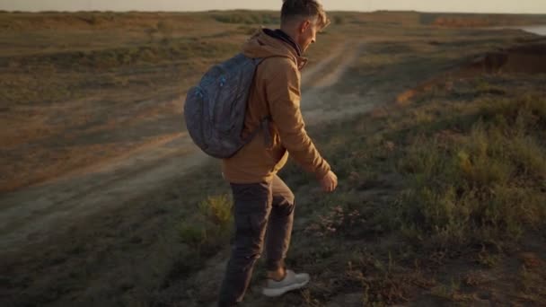 Vista laterale uomo escursionista in abbigliamento casual sale sulla costa ripida collina erbosa mare e guardando l'alba a deserta ampia spiaggia di mare — Video Stock