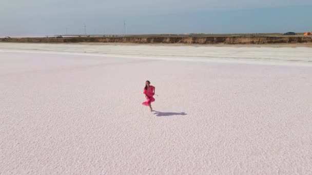 Drohnenaufnahmen der schönen fröhlichen Frau im rosafarbenen Kleid beim Joggen und Laufen über die Salzebenen des rosafarbenen Sees — Stockvideo