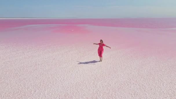 Filmische Drohnenaufnahme des farbenfrohen rosafarbenen Sees mit weißer Salzküsten-Paradieslandschaft. Mädchen in wehendem rosa Kleid spaziert an atemberaubender Küste — Stockvideo