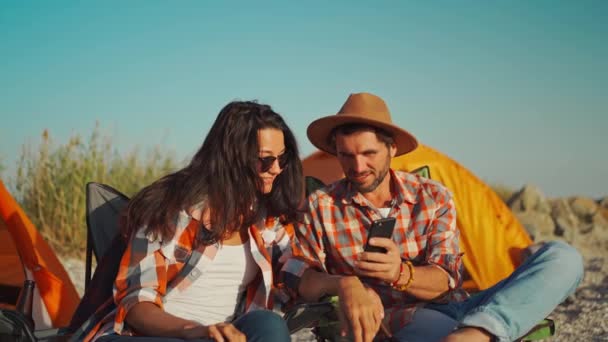Beautyful romantic couple in checkered shirts sitting at seaside in camp chairs and looking photos and videos on smartphone, mobile phone. — Stock Video