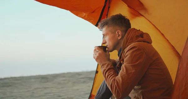Voksen mann som sitter ved teltinngangen og holder og lukter krus med kaffe i hendene – stockfoto
