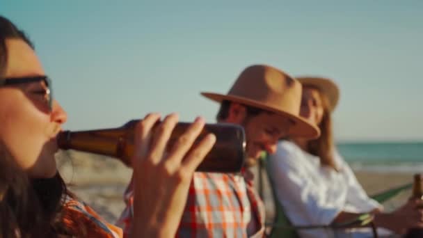 Junge, glückliche Freundinnen trinken Bier, sitzen und entspannen auf Liegestühlen am Strand und hören dem schönen Mann zu, der an einem Sommerabend bei Sonnenuntergang Gitarre spielt — Stockvideo