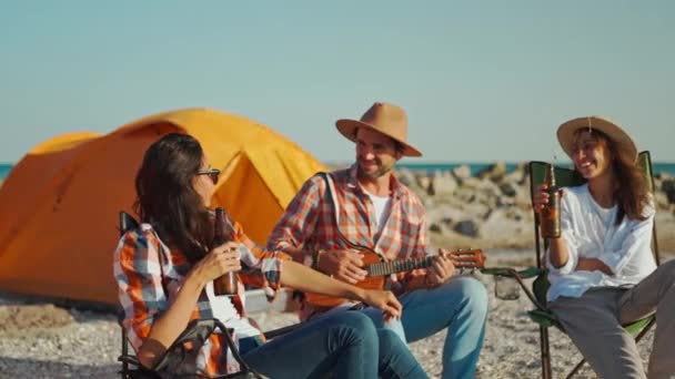 Amigos relajándose en la playa. guapo hombre en sombrero jugando ukelele y dos hermosas novias divirtiéndose, relajándose en sillas de campamento y bebiendo cerveza — Vídeo de stock