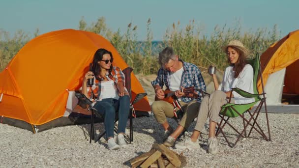 Disparo de cuerpo entero: tres mejores amigos se sientan en la playa cerca de tiendas de campaña y se divierten juntos hablando, riendo y jugando ukelele — Vídeos de Stock