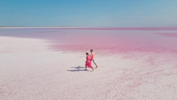 Повітряний епічний дрон зображення прекрасної романтичної пари, що йде по яскравій барвистій рожевій воді мінерального озера з білим солоним узбережжям — стокове відео