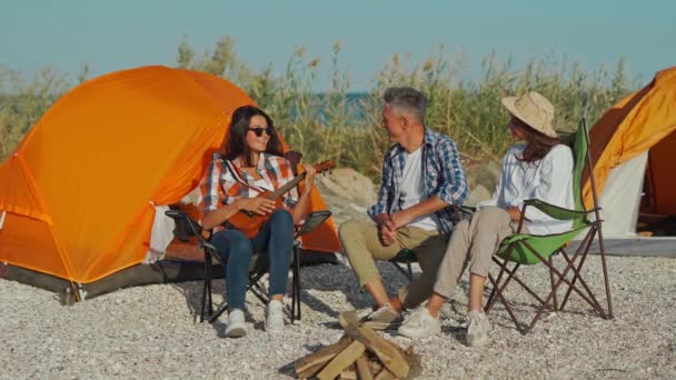 Bela asiático jovem mulher em óculos de sol e xadrez camisa jogar ukulele e casal amigos ouvindo e se divertindo, relaxando no acampamento cadeiras — Vídeo de Stock