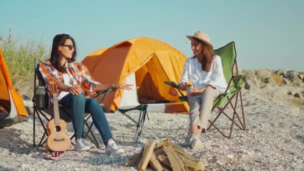 Pareja encantadora mujeres hermosas novias sentadas en sillas de camping en la costa del mar, divertirse hablando y riendo. chicas relajándose en el campamento con tiendas de campaña en la playa al atardecer — Vídeos de Stock