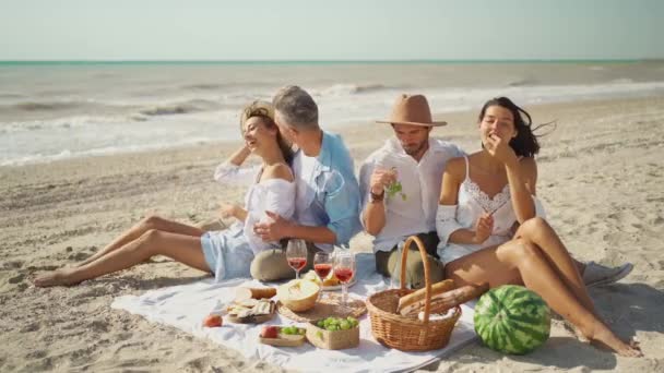 Concepto de picnic de amistad, ocio y playa - grupo de amigos felices comiendo frutas en el picnic en la playa en verano — Vídeo de stock