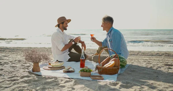 欧洲快乐的同性恋夫妇在海滩喝酒，享受浪漫的野餐 图库照片