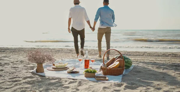 Homofile par som går på stranden, fokuserer på piknikpledd med vin, glass og mat stockbilde