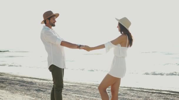 Liebendes junges Paar mit Hüten, die einander in die Augen starren und sich küssen, während sie an einem sonnigen Morgen Hand in Hand an einem Sandstrand stehen — Stockvideo