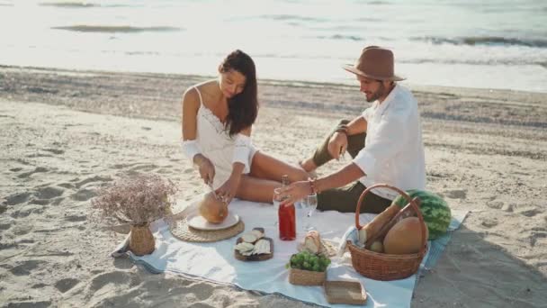 Belo casal tomando café da manhã romântico com muita comida saborosa e vinho, sentado em manta piquenique na praia com vista para o mar — Vídeo de Stock