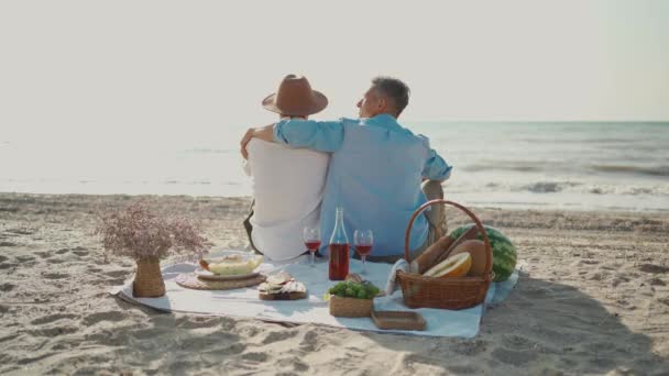 Гомосексуальная пара, гомосексуалисты устраивают пикник на пляже, сидят на одеяле и обнимаются, смотрят друг на друга с любовью — стоковое видео