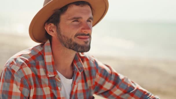 Jóképű elegáns alkalmi férfi utazó kockás ing és kalap ül a tengerparton, és élvezi a békés természet, a szabadság és a nyugalom — Stock videók