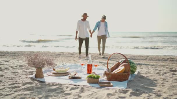 Silhouette gay couple walking by sea beach, focus on picnic blanket with wine, glasses and food. — Stock Video