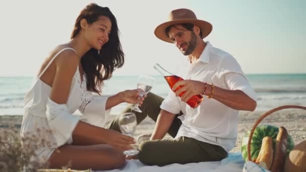 Romântico jovem casal beber vinho e desfrutar de piquenique na praia, homem derramando videira rosa em copos. — Vídeo de Stock