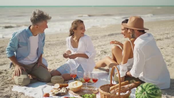 Cuatro amigos milenarios tienen picnic en la playa de arena en el día soleado siguiente mar ondulante — Vídeo de stock