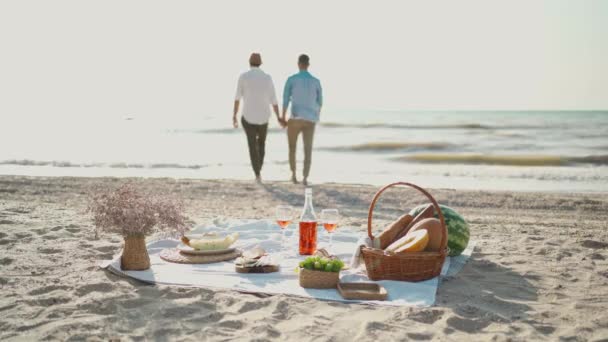 Силует гей-пара, що йде на морському пляжі, зосередитися на пікніку ковдру з вином, окулярами та їжею . — стокове відео