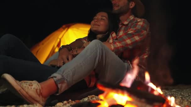 Tierna pareja romántica sentada en el fuego en camping. hombre abrazando a mujer delante de la hoguera en la fiesta de playa en la oscuridad — Vídeo de stock