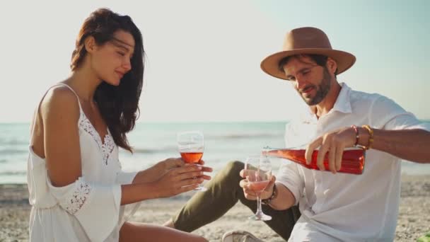 Casal em desgaste branco ter ao ar livre namoro, piquenique com vinho, sentado em cobertor na praia de areia com vista para o mar. homem derramando vinho rosa em copos e casal torcendo. — Vídeo de Stock