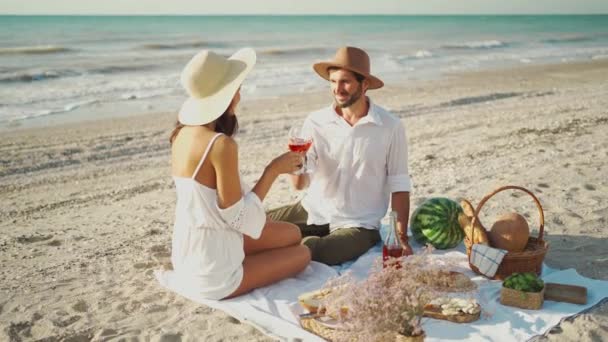 Attraktives Paar in Hüten beim romantischen Picknick mit viel leckerem Essen und Wein, auf Picknickdecke am Strand mit Meerblick sitzend. — Stockvideo