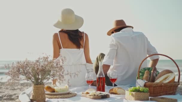 Vue arrière D'heureux couple romantique en chapeaux sur pique-nique de plage. joyeux homme et femme assis sur la couverture de pique-nique que debout et courir à la mer — Video