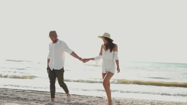 Şapkalı romantik bir çift kumsalda el ele yürüyorlar. Birbirlerinin gözlerine bakıp el ele öpüşüyorlar. — Stok video