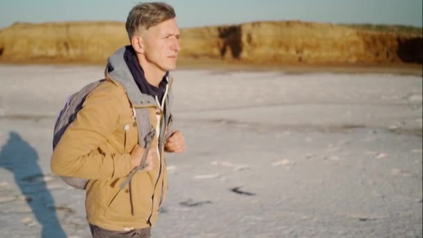 Медленный кадр молодого человека, идущего по берегу озера с белыми солеными квартирами и крутым глиняным берегом — стоковое видео