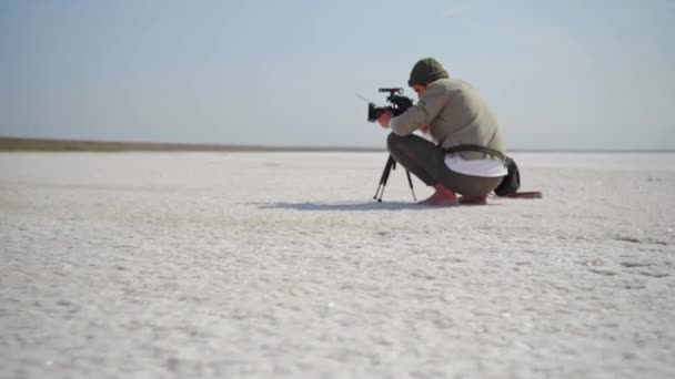 Kameramozgás az operatőr körül a munkahelyen. férfi filmezés segítségével kamera állvány ül fehér sós strand száraz ásványi tó, mint egy sivatag — Stock videók