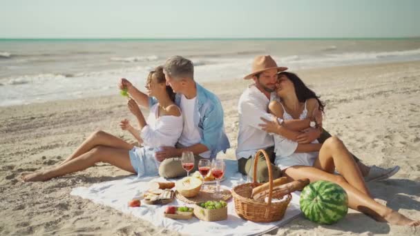 Vänskap, fritid och strandpicknick koncept - grupp glada vänner äter frukt på picknick på stranden på sommaren — Stockvideo