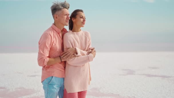 Hombre cariñoso abrazando suavemente a la joven atractiva mujer en suéter rosa en la costa salada blanca del lago rosa parece desierto. — Vídeo de stock