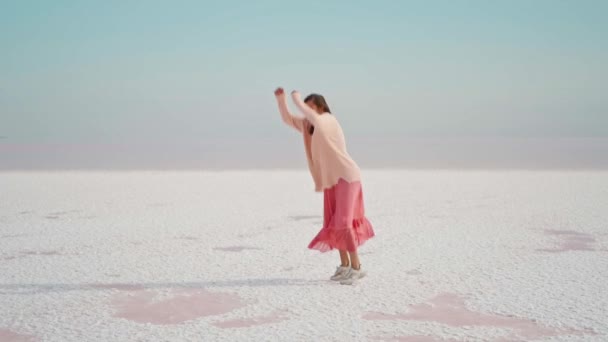 분홍색지나치게 커 보이는 옷을 입고 하얀 사막 배경에 발을 들고 우스꽝스럽게 점프하는 행복 한 미소짓는 여인 — 비디오