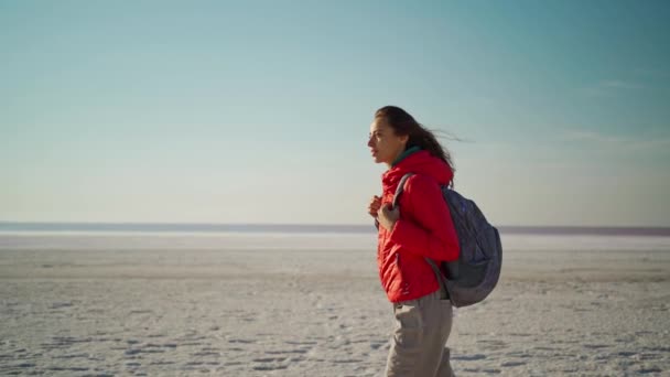 Slow motion wandelende vrouw in rode jas en rugzak op wit zout strand — Stockvideo