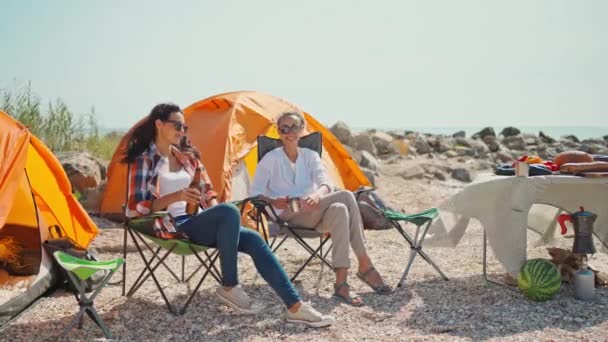 Två vackra avslappnade flickvänner resenärer njuter av sommarsemester och dricka kall öl på varm solig dag i camping på havskusten — Stockvideo