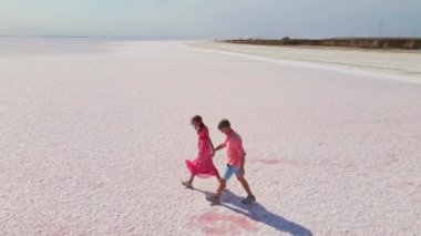 Pembe elbiseli mutlu çiftin parlak pembe mineral gölünün beyaz kıyısında eğlenirken ve neşe içinde koşarken çekilmiş hava aracı görüntüleri.